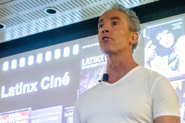 Frederick Luis Aldama speaks in front of a large screen displaying Latinx movie posters